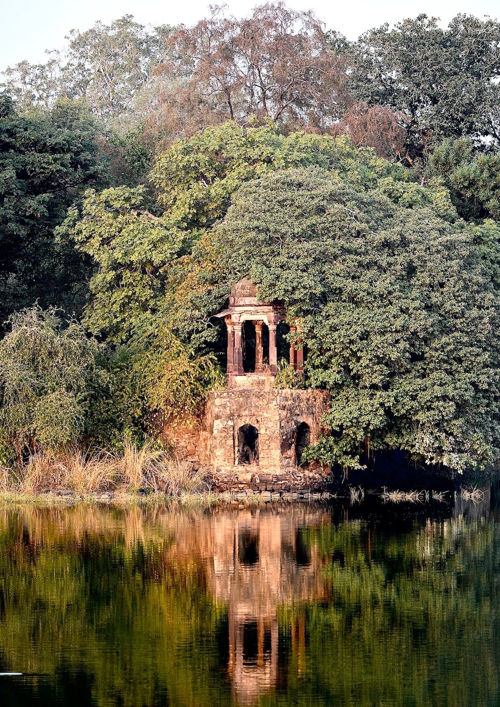 An old stone pavilion ruin among trees, mirrored in the still water of a lake